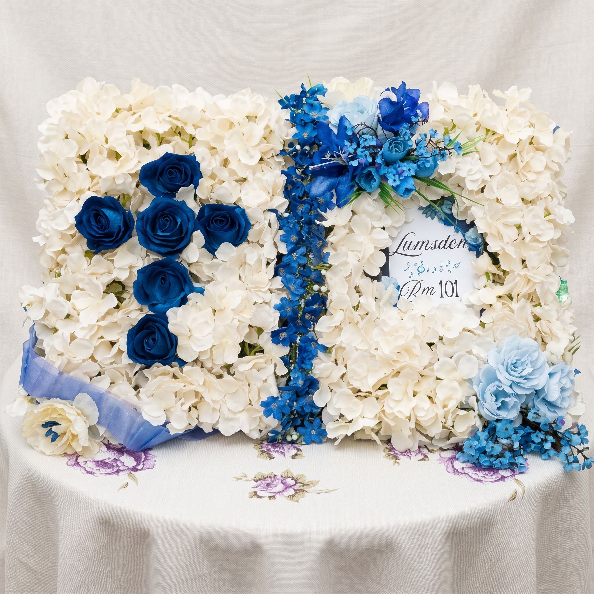 Open book arrangement of cream hydrangeas with a cross of royal blue roses on the left page, a trail of blue delphiniums down the centre, pale blue roses in the corner, and a small name plaque on the right page.
