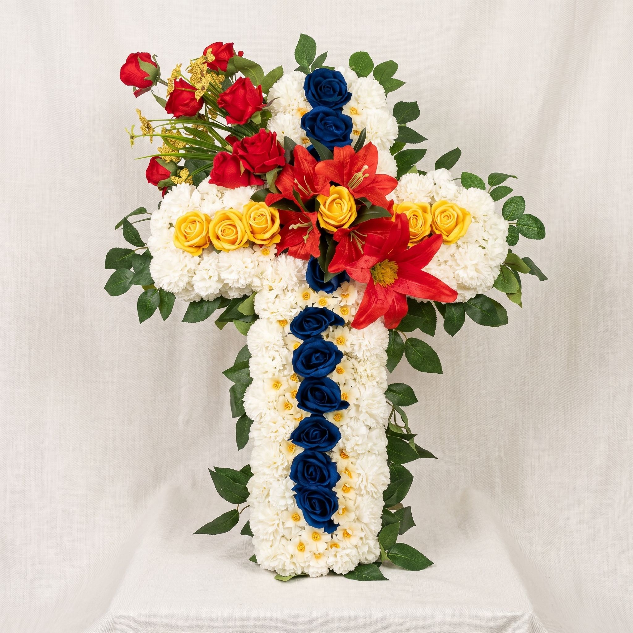 Standing cross of white carnations with blue roses down the centre, yellow roses across the arms, and red lilies and roses at the top.