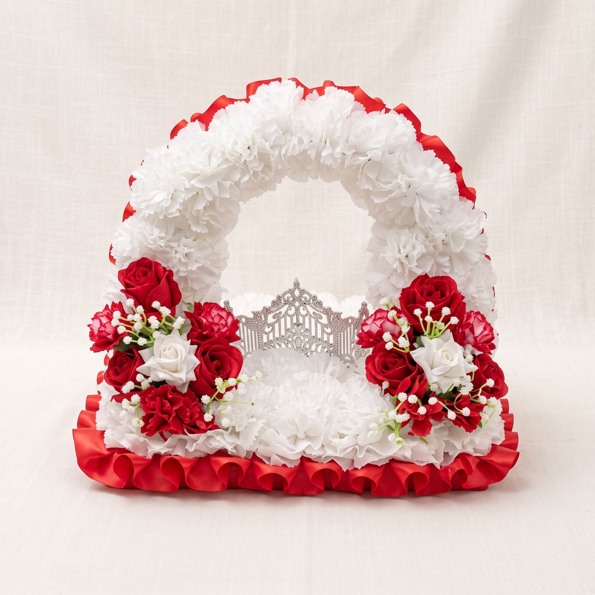 Arched arrangement of white carnations with red satin ruffle trim, red and white roses on each side, and a silver gate ornament in the centre.