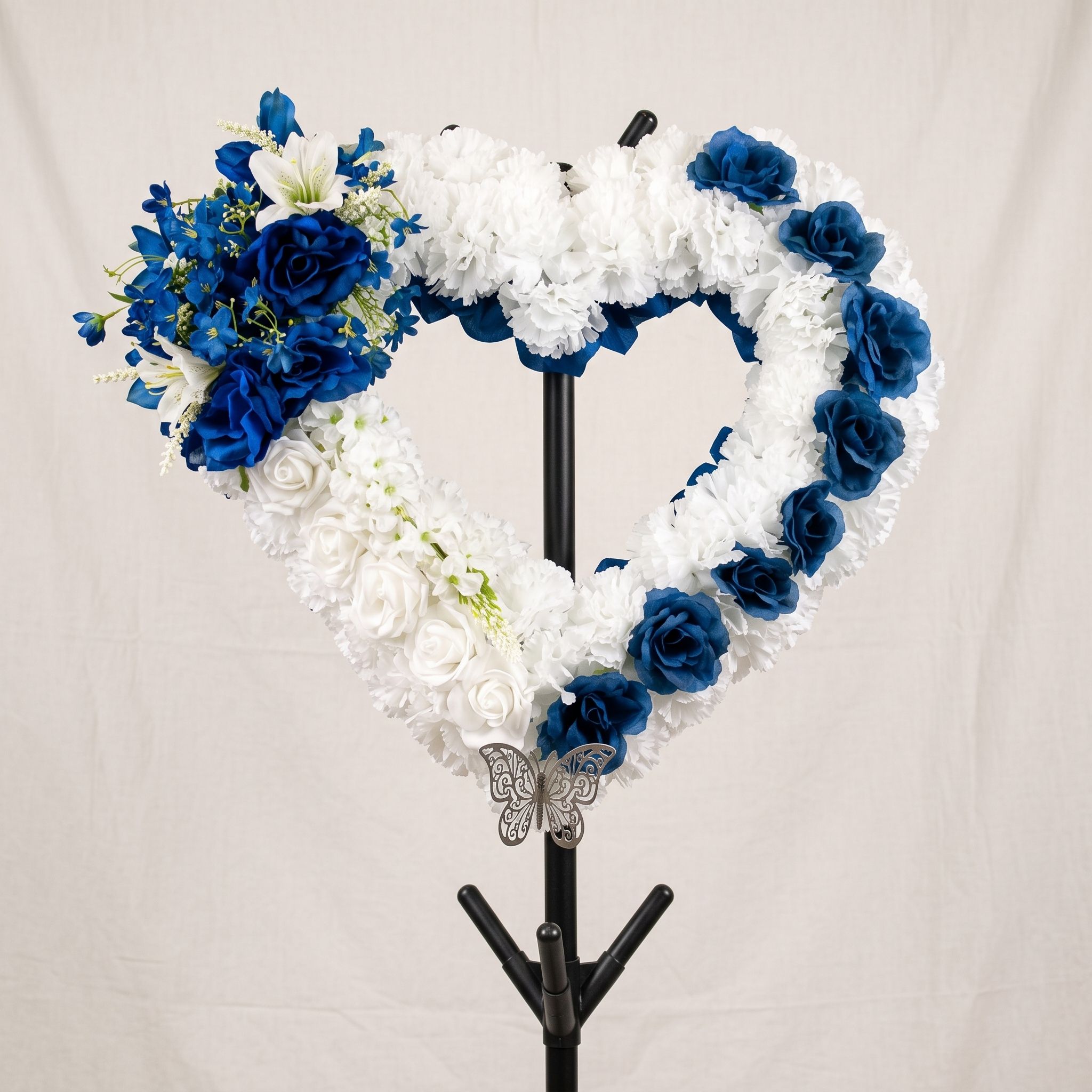 Open heart wreath of white carnations and navy blue roses on a black stand, with a blue and white spray at the top left and a silver butterfly at the bottom.