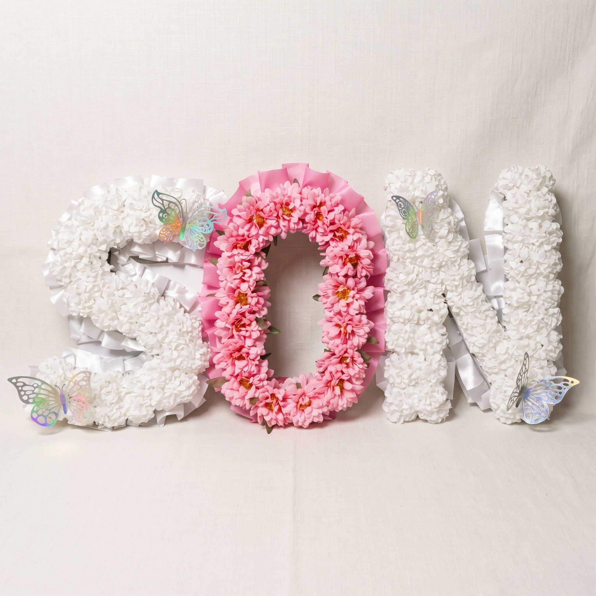 The word "SON" in flower letters: the S and N in white carnations with white ribbon edging, the O in pink chrysanthemums with pink ribbon, and four iridescent butterflies.