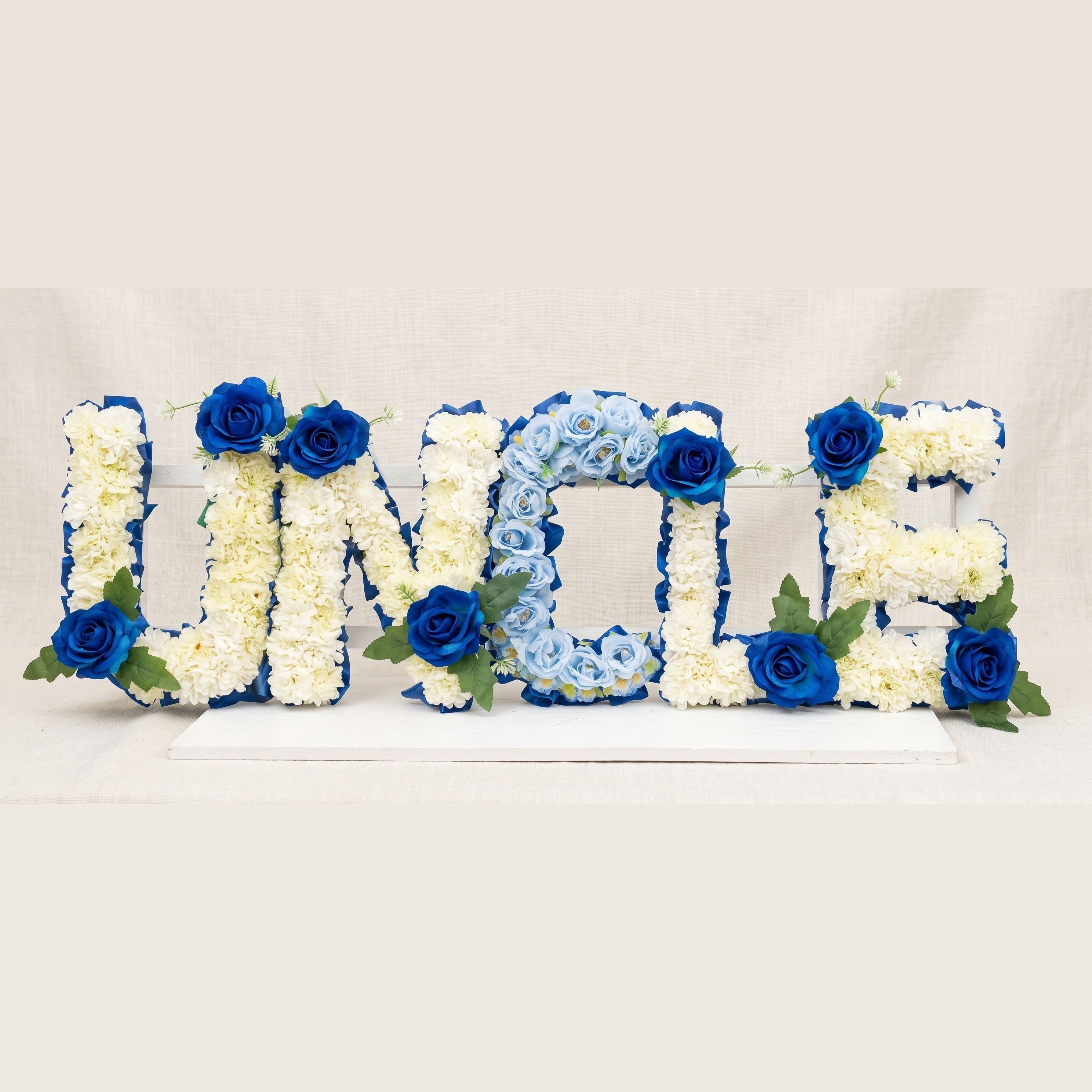 The word "UNCLE" in standing flower letters: four letters in cream chrysanthemums and the C in pale blue roses, each edged in royal blue ribbon and dressed with royal blue roses.
