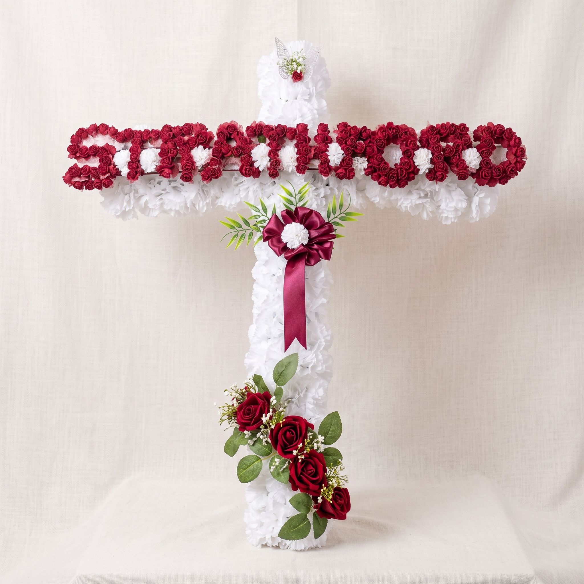 Cross of white carnations with a name spelled in small red roses across the arms, a burgundy ribbon rosette, and red roses near the base.