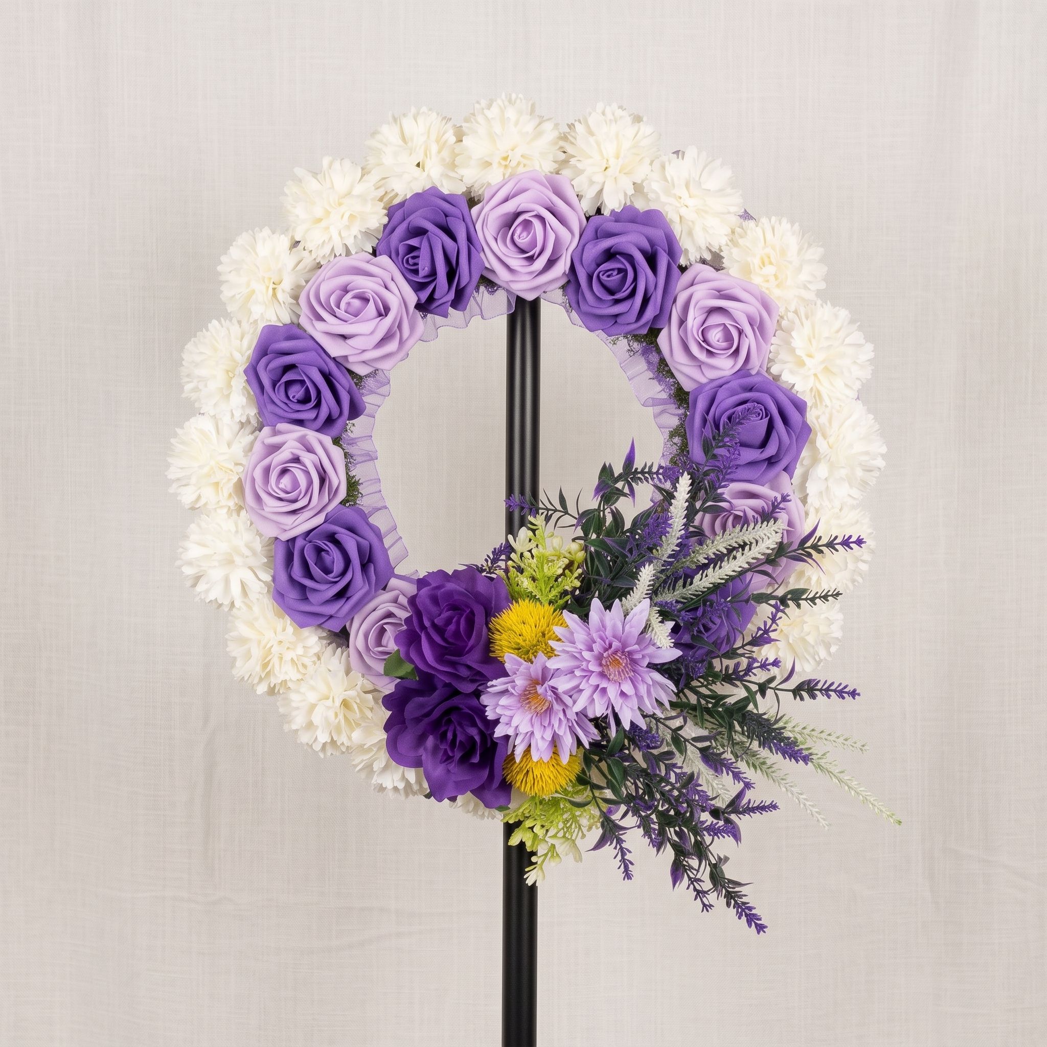 Round wreath on a black stand, with an outer ring of white carnations, an inner ring of purple and lilac roses, and a spray of lilac chrysanthemums, yellow blooms and lavender at the lower right.