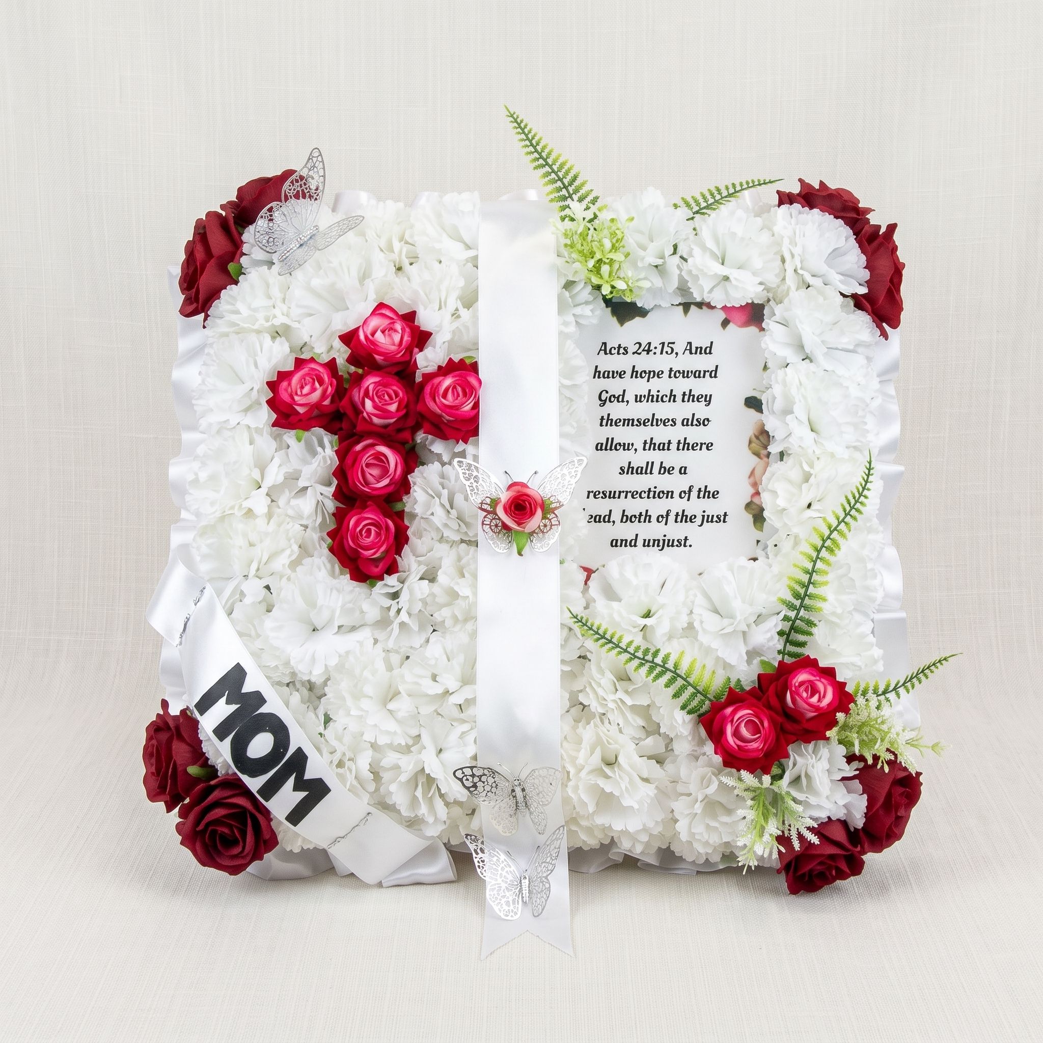 Square pillow of white carnations with a red rose cross, a framed Bible verse, silver butterflies, and a white sash reading "MOM."