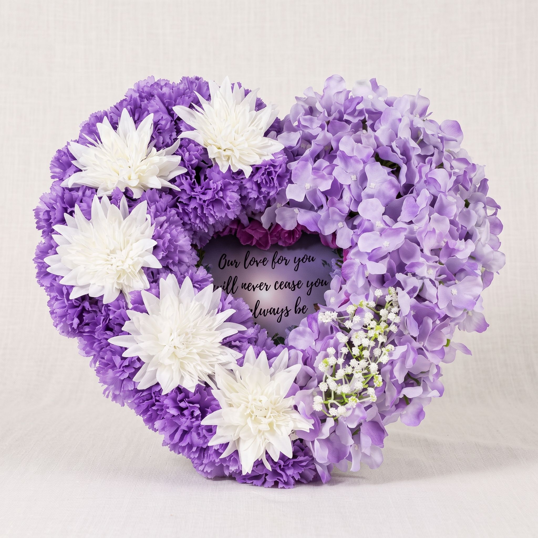 Heart of purple carnations and white chrysanthemums on the left and lilac hydrangeas on the right, with baby's breath and a small panel in the centre that reads "Our love for you will never cease, you will always be".