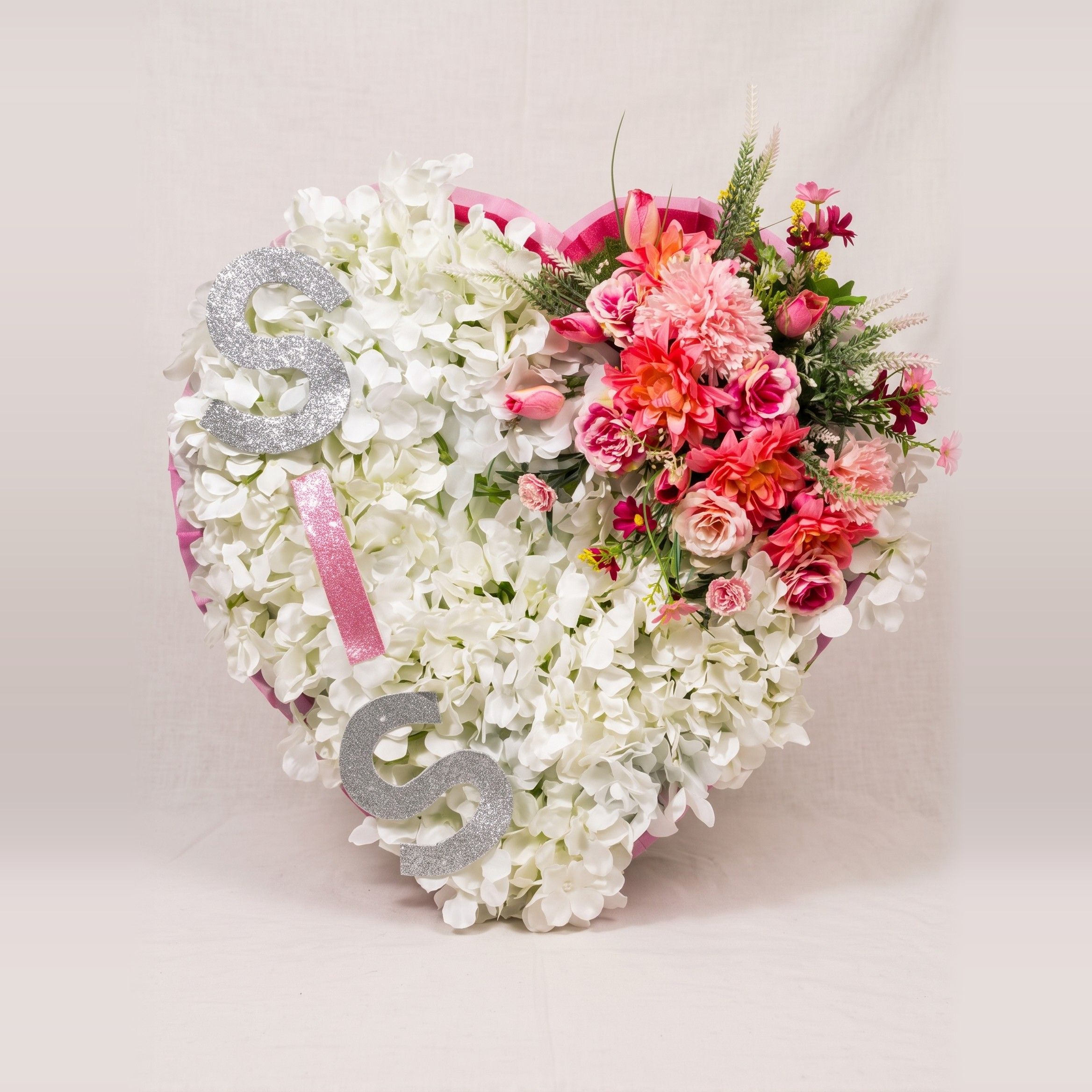 Heart of white hydrangeas edged in pink, with a spray of pink roses, coral dahlias and greenery at the top right, and silver glitter letters spelling "SIS" down the left side.